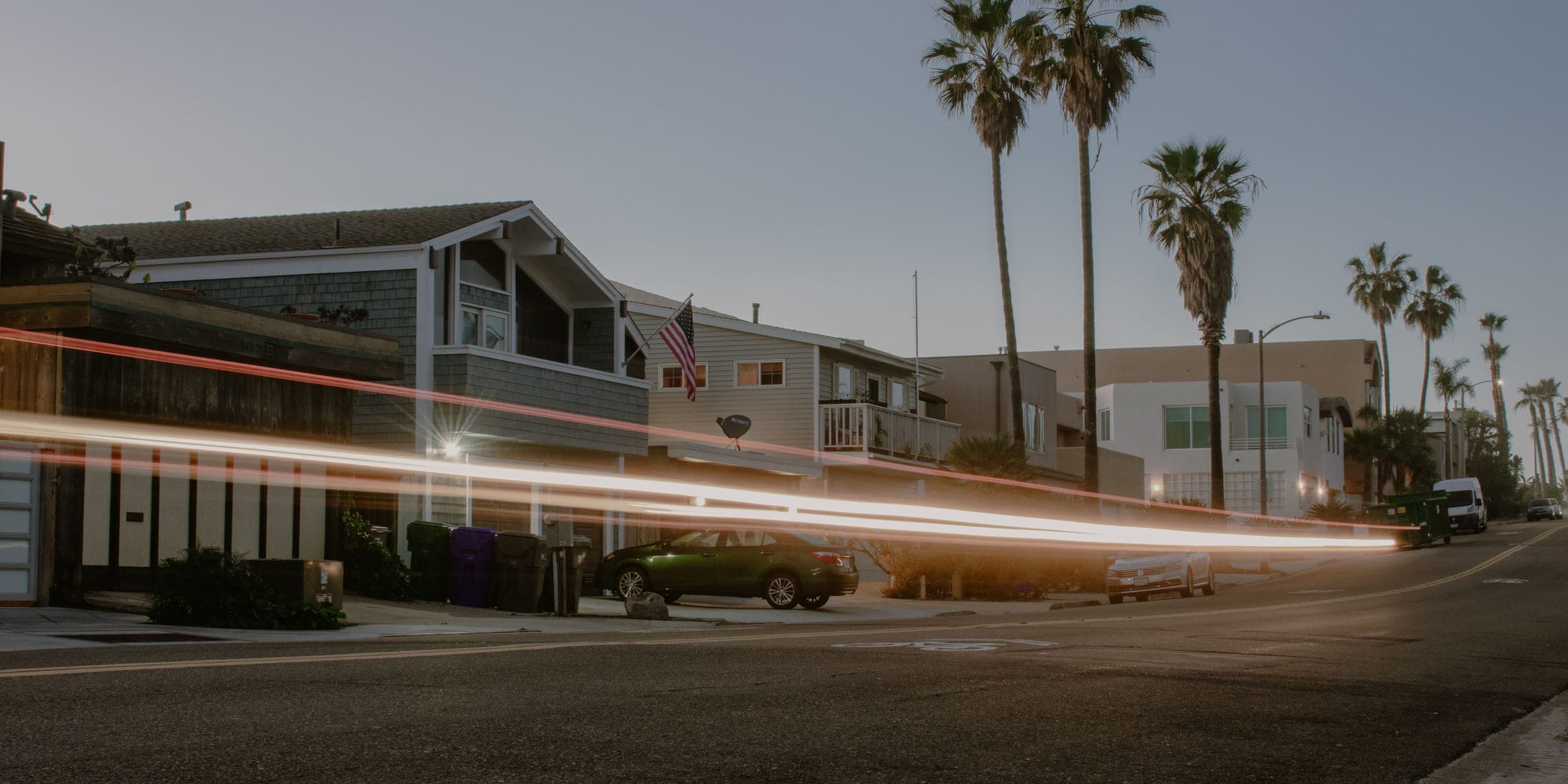 Street at dusk with shingled two-story homes, tall palm trees and car light trails