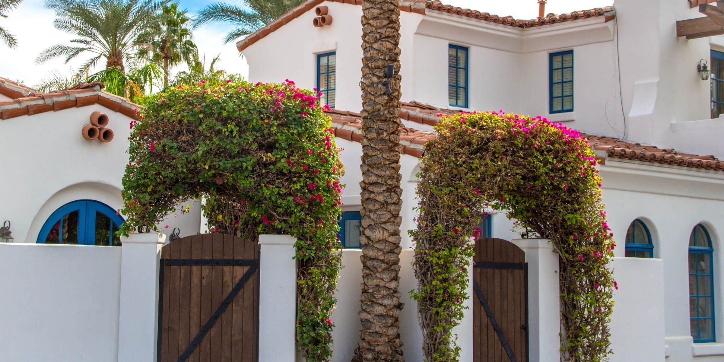 White Spanish style house with wooden gates under bougainvillea arches and palm trees