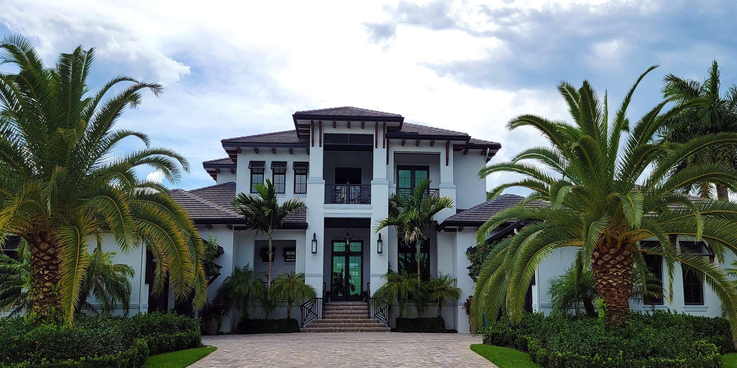 White two-story house with a dark tile roof, paver driveway and palm trees