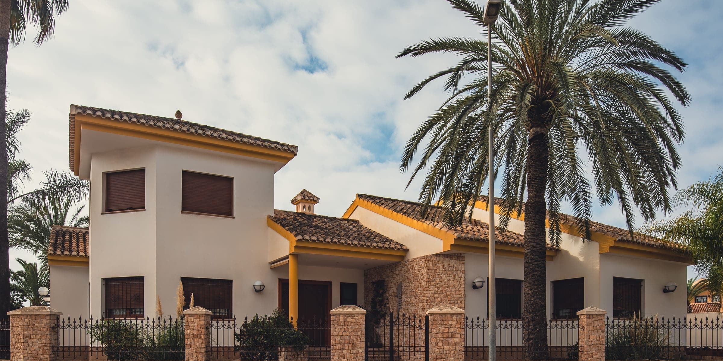 Cream stucco house with tile roofs behind an iron fence, with a large palm tree
