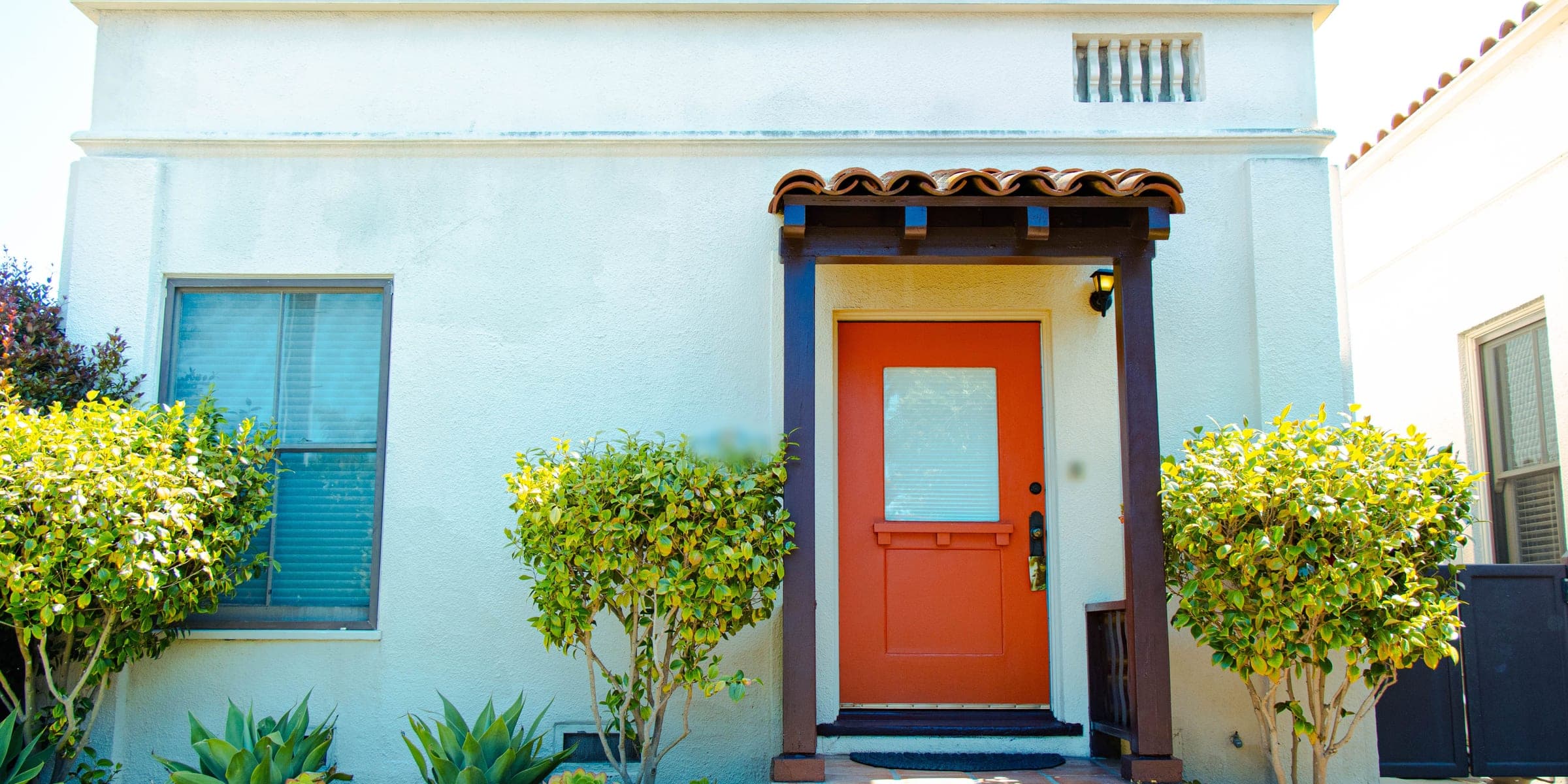 White stucco cottage with a clay tile roof line, a tiled entry porch, trimmed shrubs and succulents