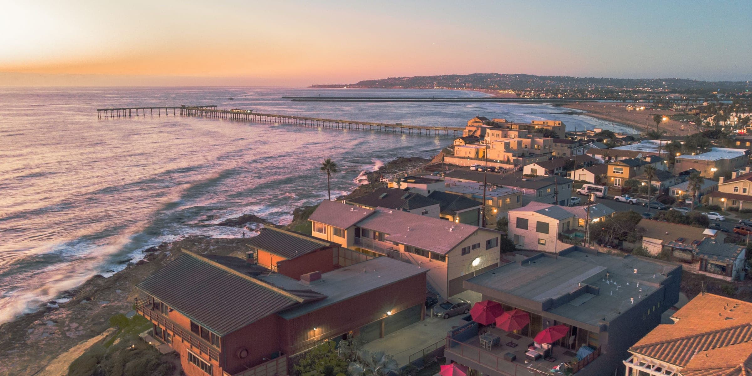 Clifftop coastal homes above the ocean with a long pier at sunset