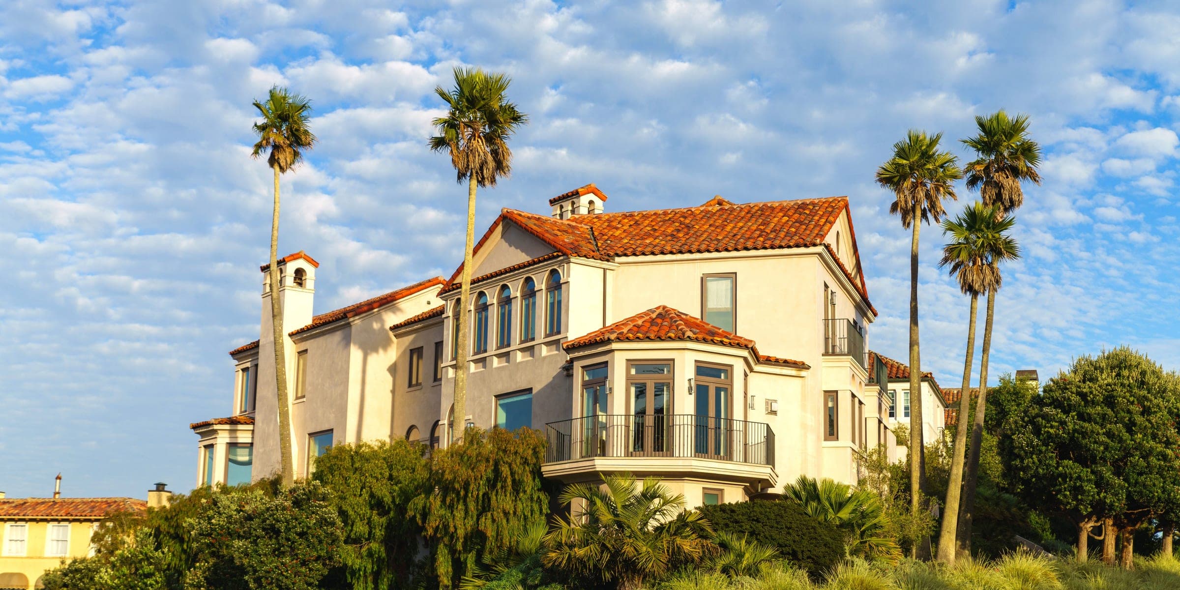 Cream Mediterranean style house with a clay tile roof, tall palms and drought-tolerant plants