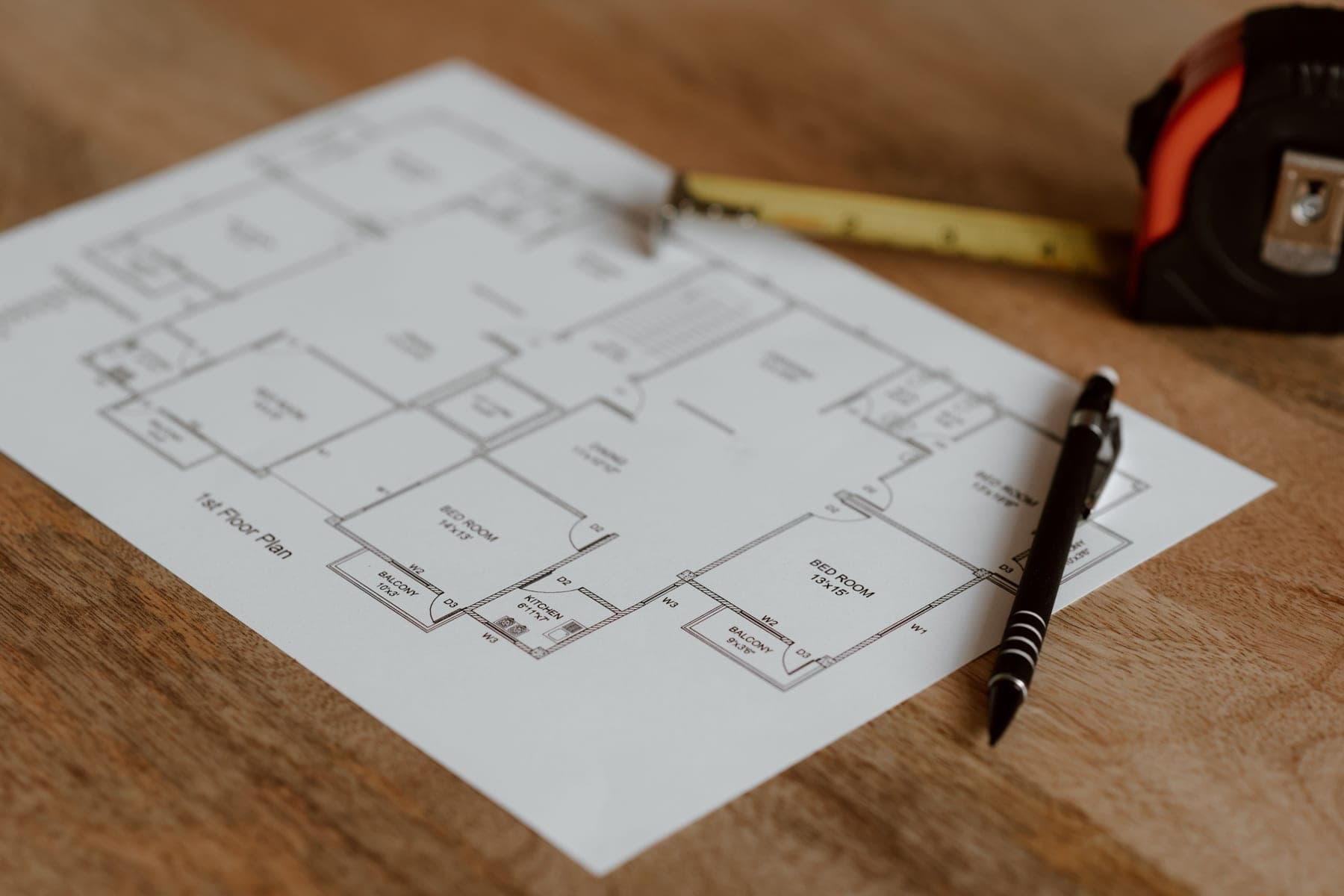 Printed single-floor house plan on a wooden table with a tape measure and a pen