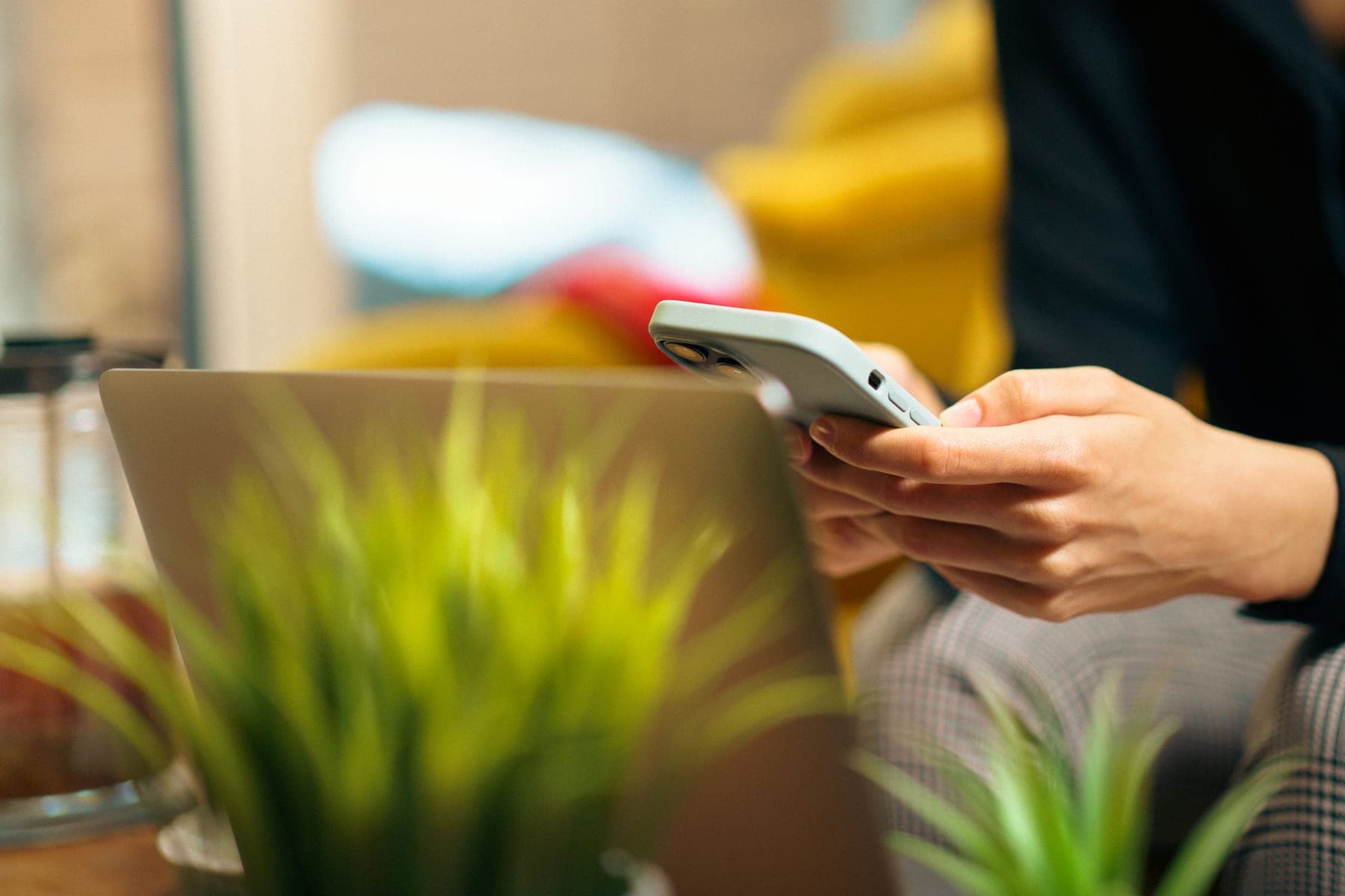 Hands holding a smartphone beside an open laptop and a houseplant