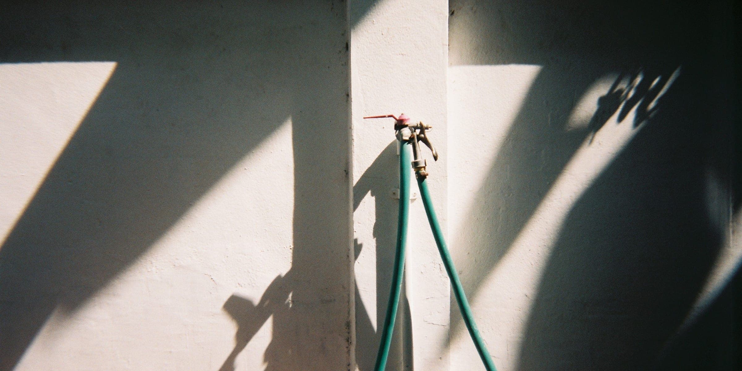 Outdoor water spigot with two green garden hoses on a sunlit white wall