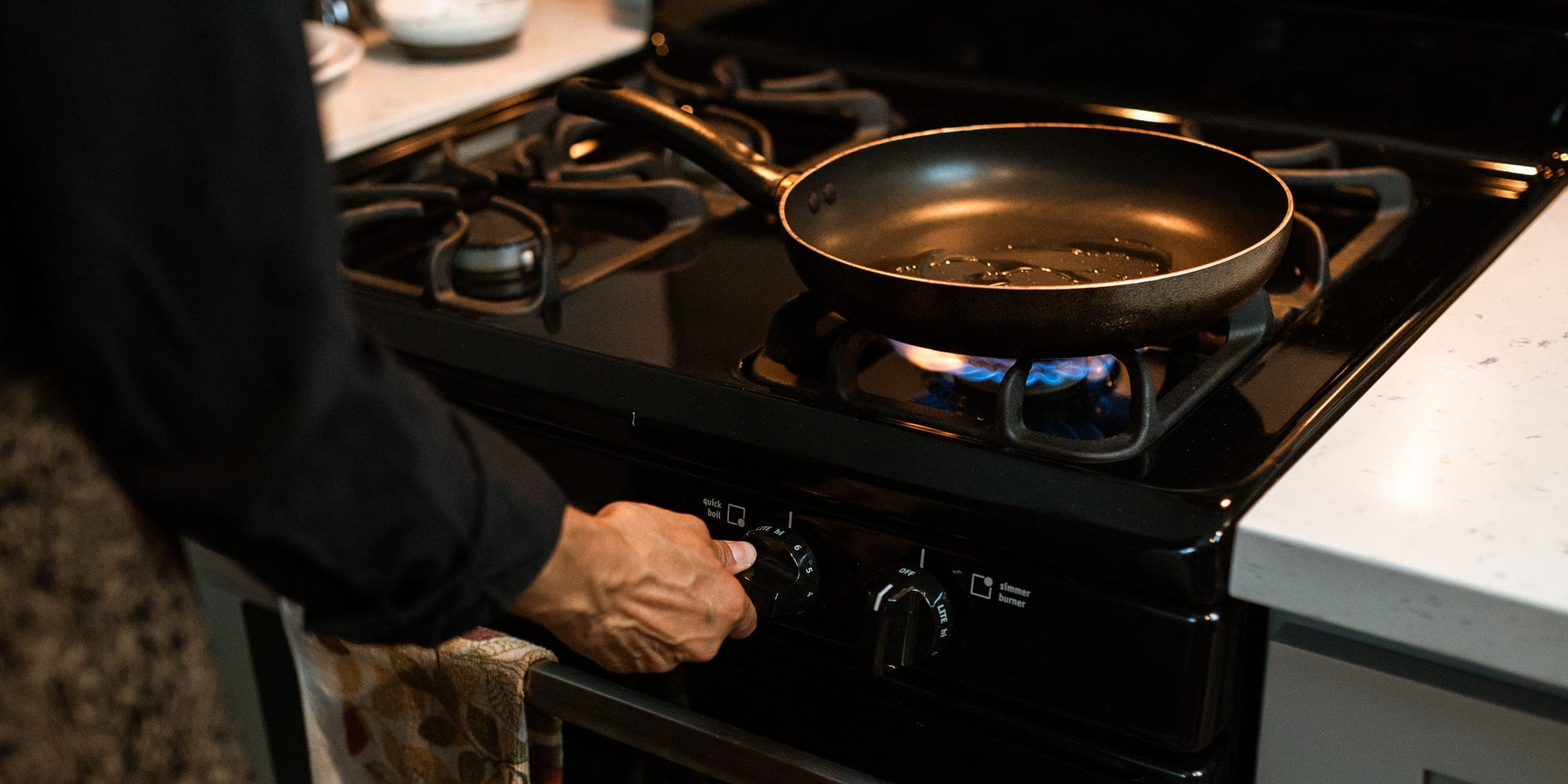 Hand turning a gas stove knob while a frying pan heats over a low flame