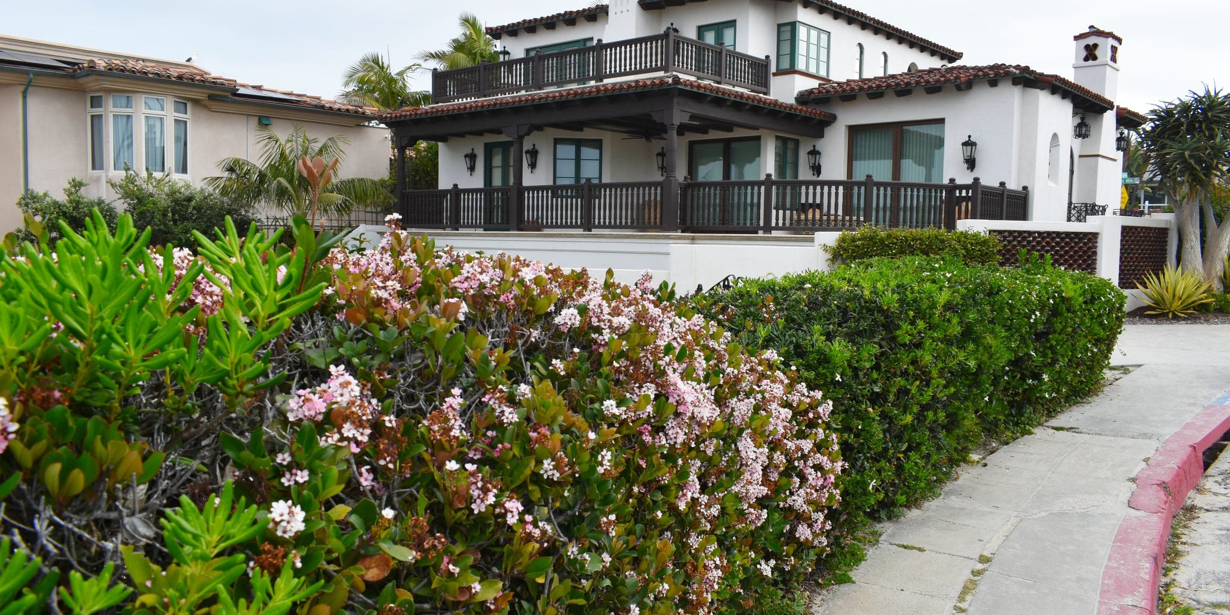 White Spanish style home with a tile roof and wood balconies above a flowering hedge