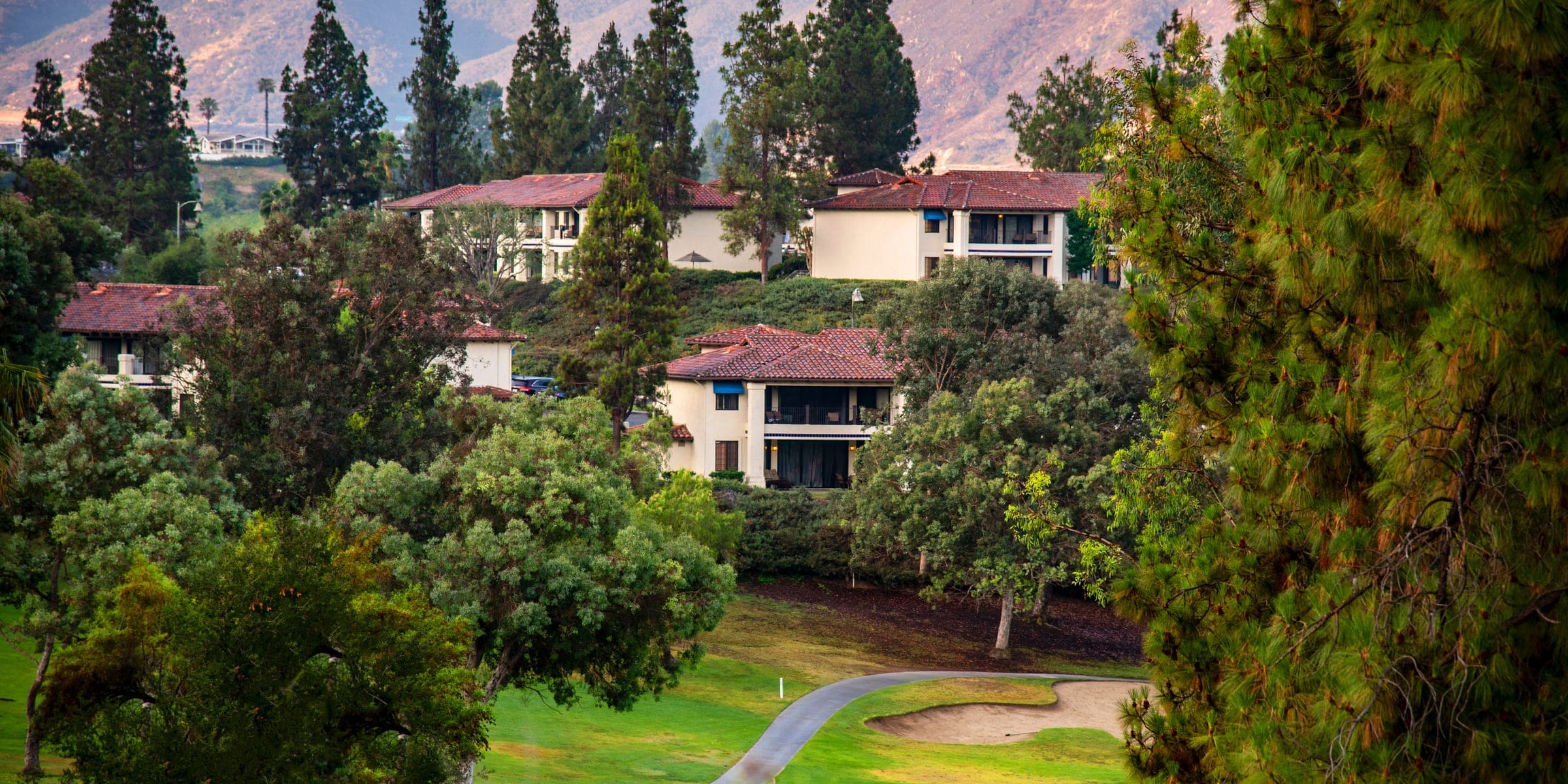 Stucco homes with tile roofs among pine trees on a hillside with mountains behind