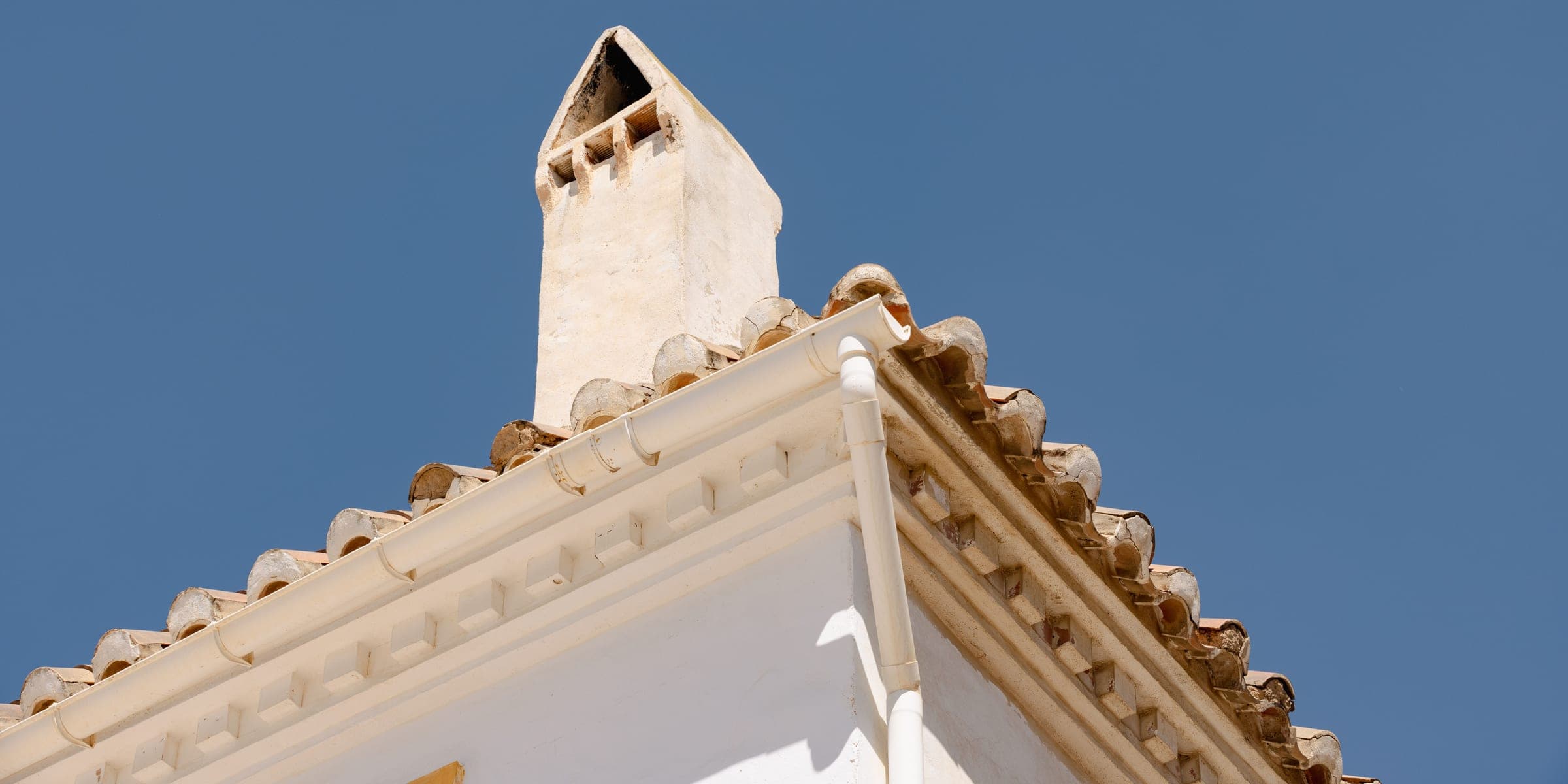 White rain gutter and downspout along the edge of a clay tile roof under a blue sky